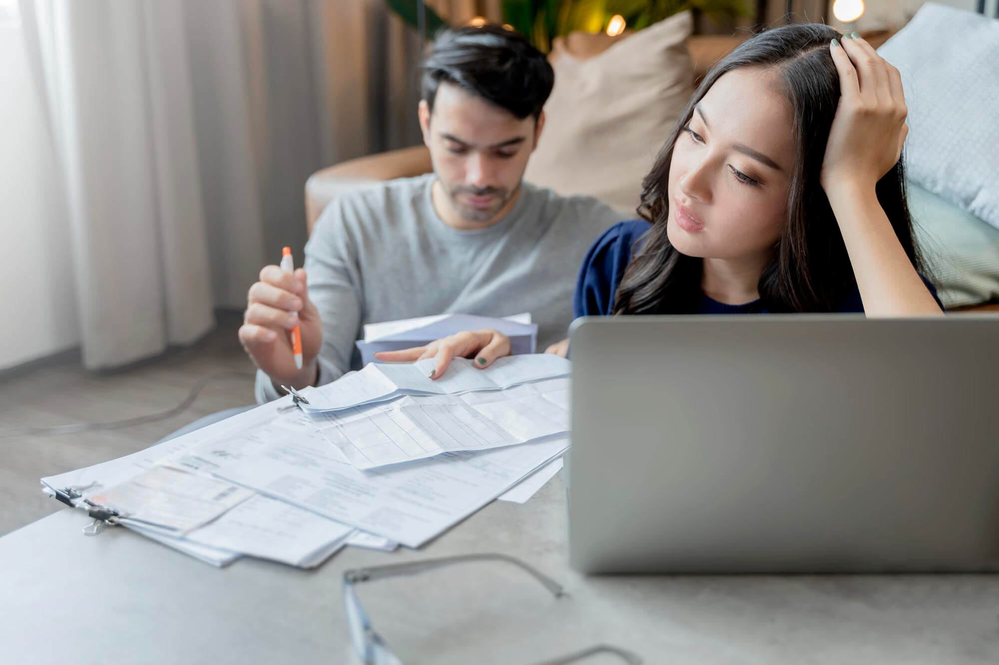 casal sentado em uma mesa, mulher asiática e homem branco os dois olhando contas e o computador parecendo confusos pensando como organizar as finanças