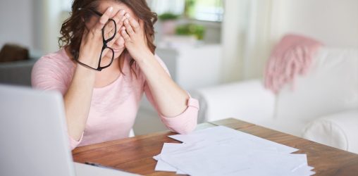 mulher sentada em uma mesa em frente a um computador e vários papéis com a mão no rosto e segurando os óculos com um semblante preocupado