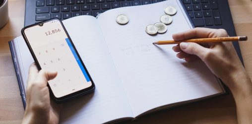 zoom de uma agenda com uma pessoa com o celular aberto na calculadora e algumas moedas do lado. a pessoa está fazendo contas e tentando se organizar financeiramente