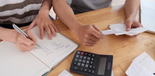 mãos de um homem e uma mulher sobre uma mesa entre papéis e calculadoras e os dois fazendo contas