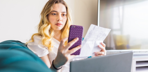 mulher loira segurando um celular e uma conta com um notebook aberto na sua frente