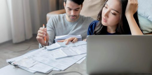 casal sentado em uma mesa, mulher asiática e homem branco os dois olhando contas e o computador parecendo confusos pensando como organizar as finanças
