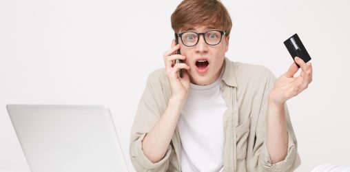 homem de cabelos curtos e óculos falando no telefone na frente do computador enquanto segura um cartão de crédito com cara de assustado