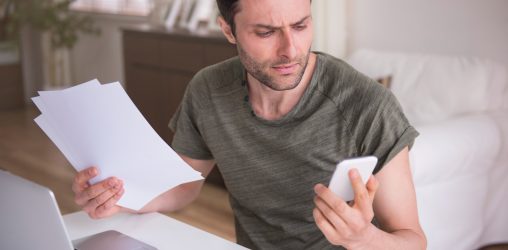 homem de camiseta verde sentado em frente a mesa com um laptop eqnaunto olha o celular com semblante preocupado e uma pilha de folhas em sua mão