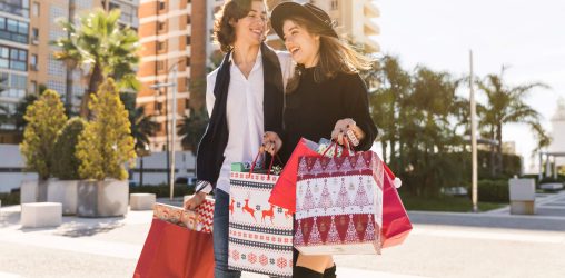 casal em frente a um prédio em uma espécie de praça segurando sacolas de natal com semblante feliz depois de fazer compras