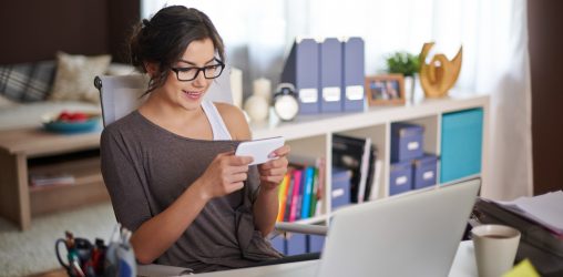 mulher de óculos e cabelo preso olhando com um rosto feliz para o celular no escritório
