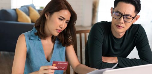 casal asiático olhando para o computador com um semblante sério enquanto checa um papel e a mulher segura um cartão de crédito