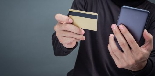 homem vestido de preto segurando um celular e um cartão na mão tentando dar um golpe