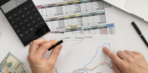 homem olhando gráficos e planinhas com uma lupa e uma calculadora do lado sobre uma mesa
