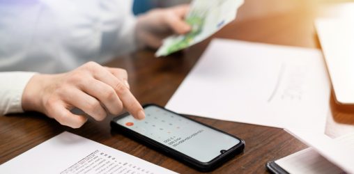 mulher lidando com finanças com a mão sobre a mesa, mexendo em um smartphone com a calculadora aberta e com algumas notas de dinheiro na mão